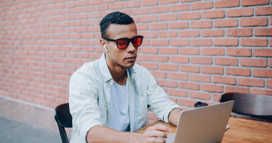 Man wearing red lens blue light blocking glasses while working on a laptop outdoors