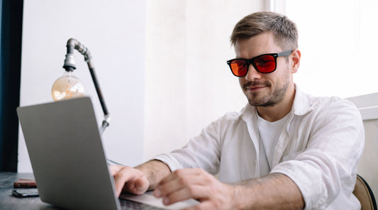 Man smiling while wearing red lens blue light blocking glasses and working on a laptop at a desk