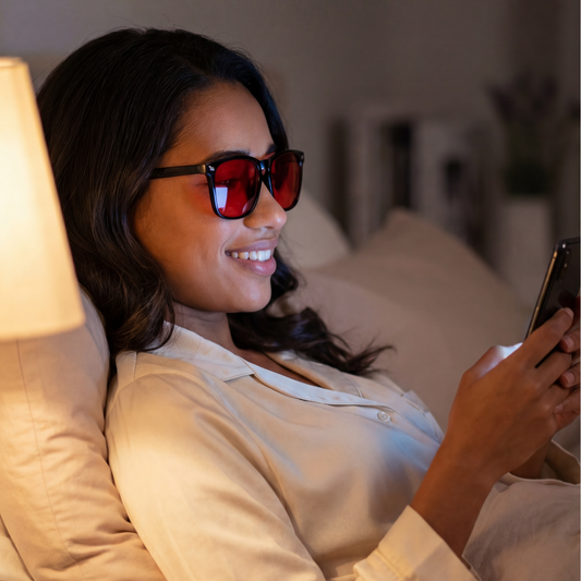 Woman wearing blue light blocking glasses with red tinted lenses while using a phone before bed in soft lamp light