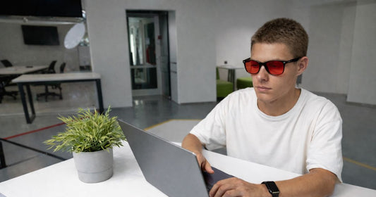 Young man wearing red lens blue light blocking glasses while working on a laptop in a modern office